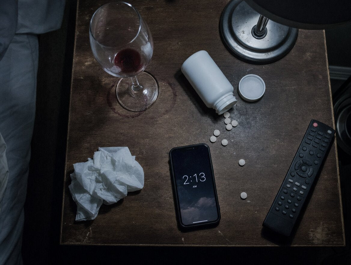 A cluttered nightstand with a wine glass, spilled tablets and a phone showing 2:13 AM